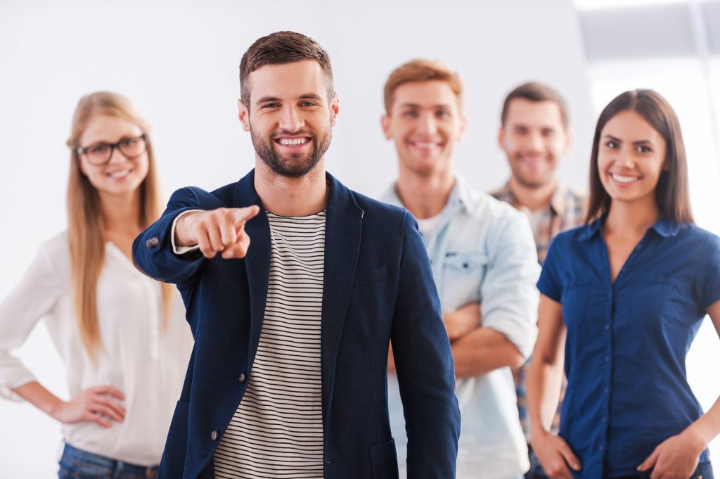 Beautiful young woman showing her thumb up and smiling while group of happy young people standing on background and smiling